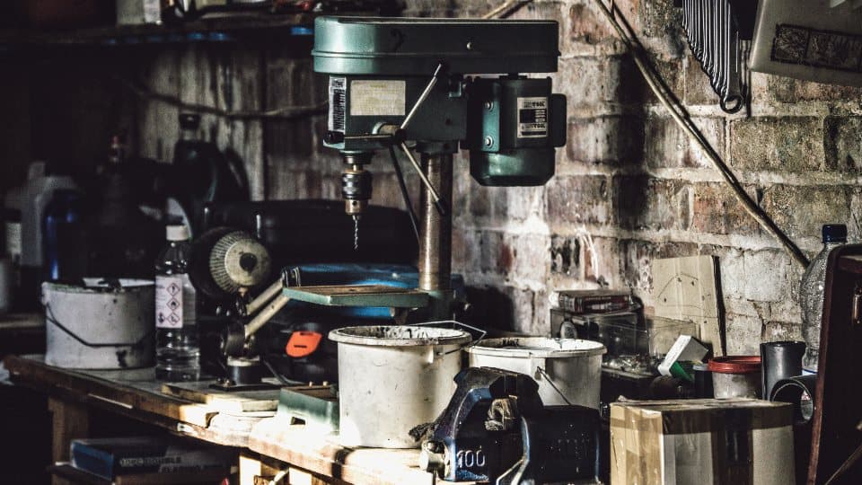 A working bench in a small workshop — a pillar drill, a vice, paint tins and tools on a brick wall